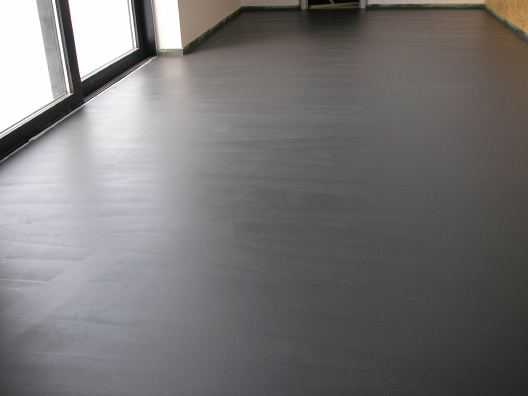 Świeżo wylana, ciemnoszara posadzka cementowa w pomieszczeniu z dużym oknem balkonowym, widoczne ślady po narzędziach wygładzających powierzchnię.